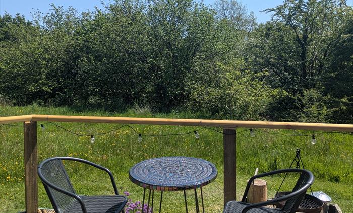 A handmade mosaic table in iridescent glass with 2 black woven chairs on the wood deck looking out to the meadow and the woodland behind.